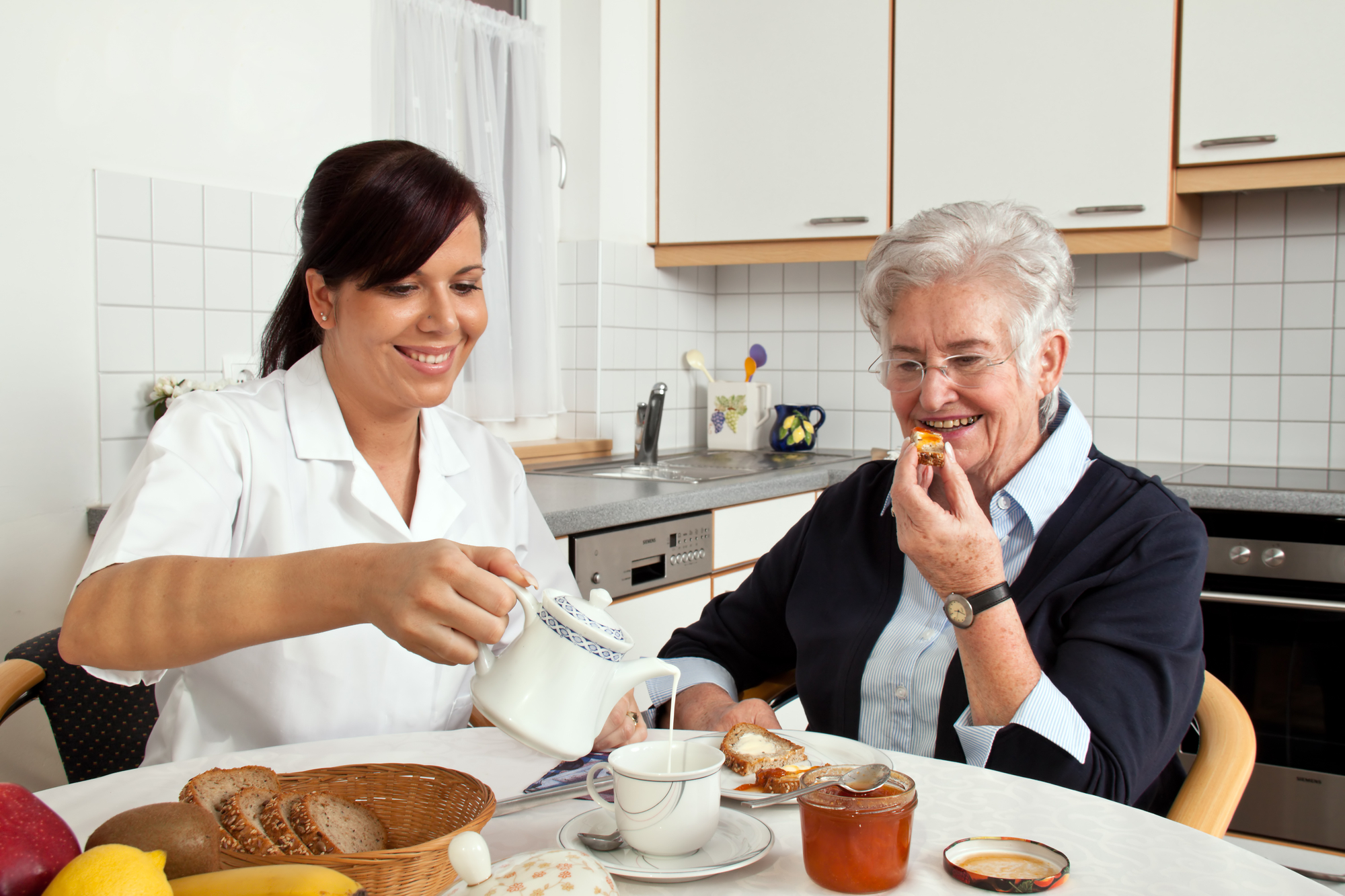 Two individuals sitting at a table in a home kitchen sharing breakfast; the younger person on the left pours cream into a teacup for a senior individual on the right, both smiling.